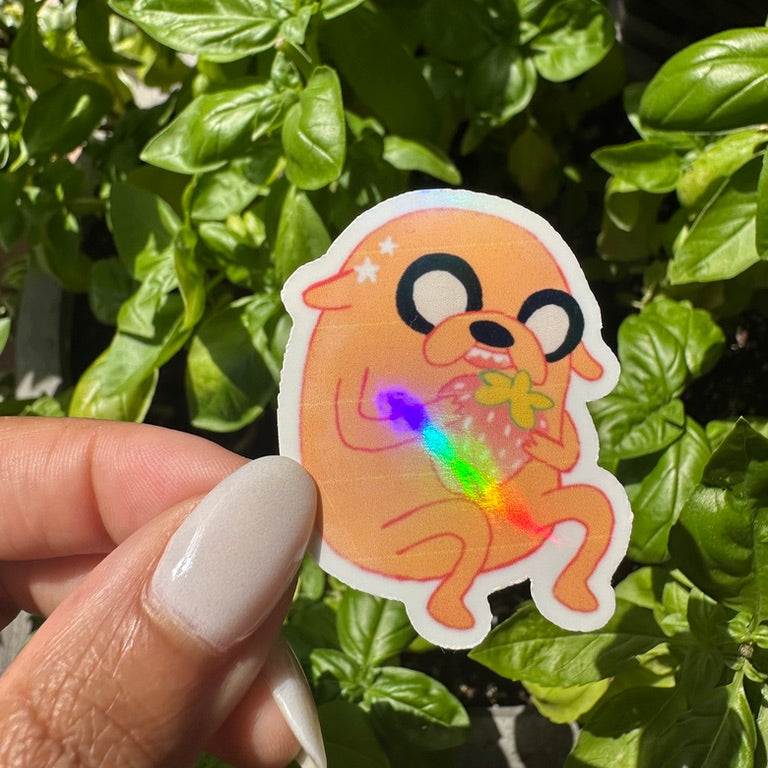 A die-cut holographic sticker of the cartoon character Jake the Dog eating a strawberry,