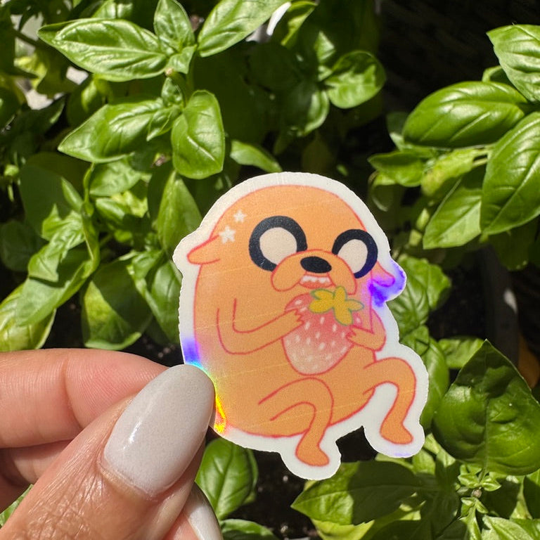 A die-cut holographic sticker of the cartoon character Jake the Dog eating a strawberry, 