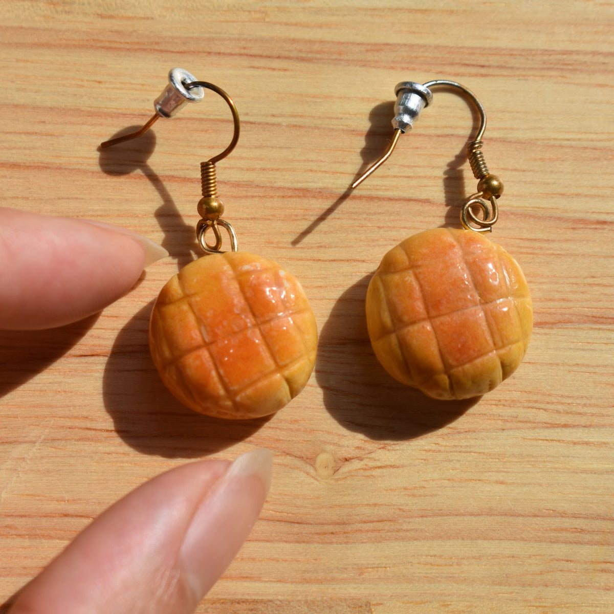 A pair of handmade earrings designed to look like melon pan bread or mini pastries, hanging from gold-toned fishhook ear wires. 