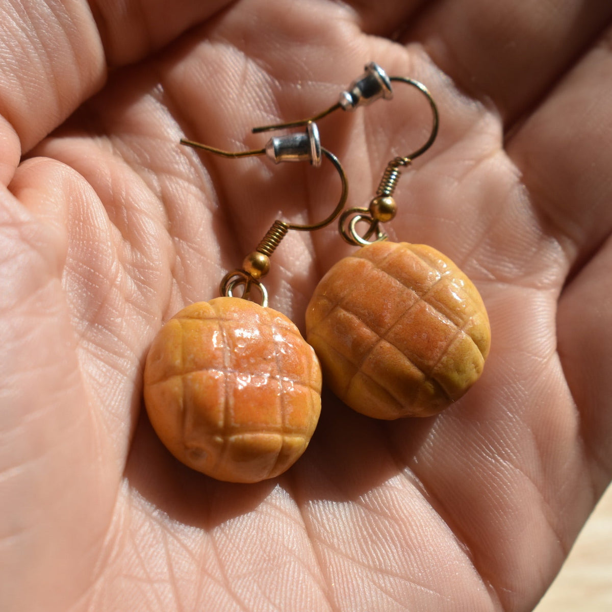 A pair of handmade earrings designed to look like melon pan bread or mini pastries, hanging from gold-toned fishhook ear wires. 