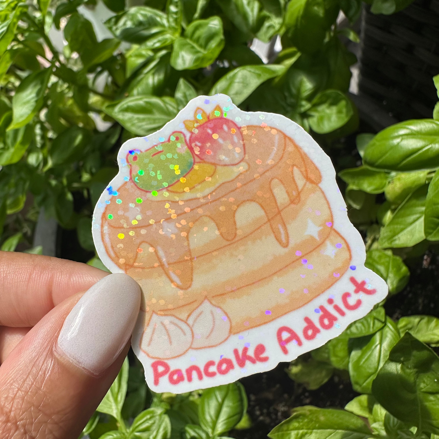 A die-cut sticker featuring a cute illustration of a layered pancake stack with a strawberry and a green frog on top, with the text 'Pancake Addict' in pink. The background is filled with green basil leaves in a sunny garden setting.