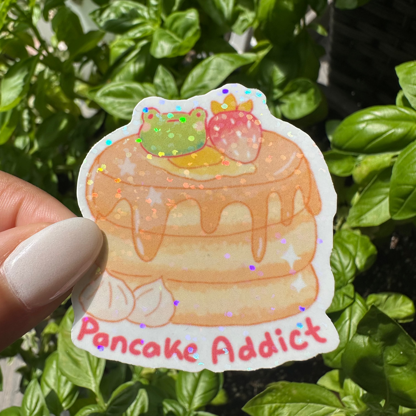 A die-cut sticker featuring a cute illustration of a layered pancake stack with a strawberry and a green frog on top, with the text 'Pancake Addict' in pink. The background is filled with green basil leaves in a sunny garden setting.