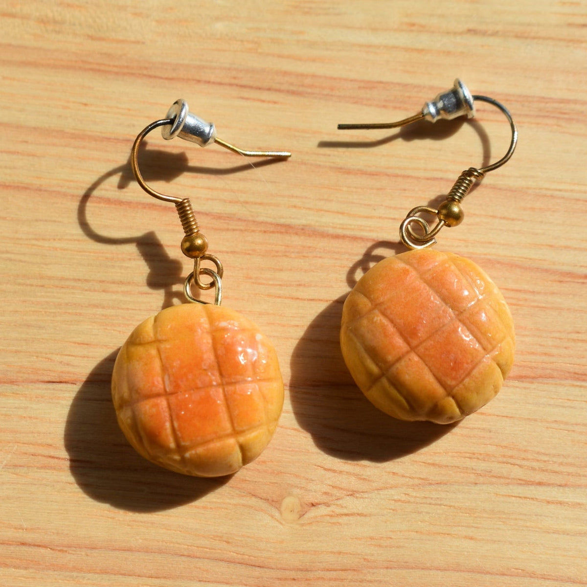 A pair of handmade earrings designed to look like melon pan bread or mini pastries, hanging from gold-toned fishhook ear wires. 