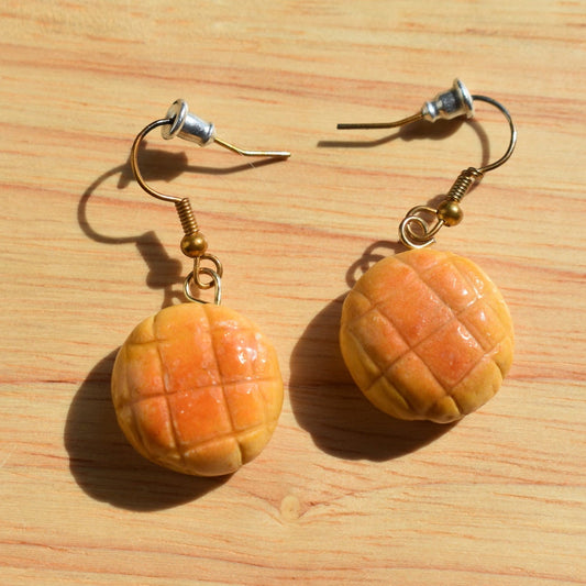 A pair of handmade earrings designed to look like melon pan bread or mini pastries, hanging from gold-toned fishhook ear wires. 
